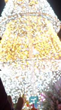 Young woman wearing a yellow beanie hat looking up at a huge, modern christmas tree made of glowing white and gold string lights in a city square during a festive winter evening