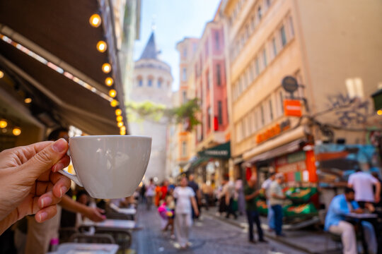 Hand holding a white coffee cup in a bustling street with colorful buildings and the Galata Tower visible in the background