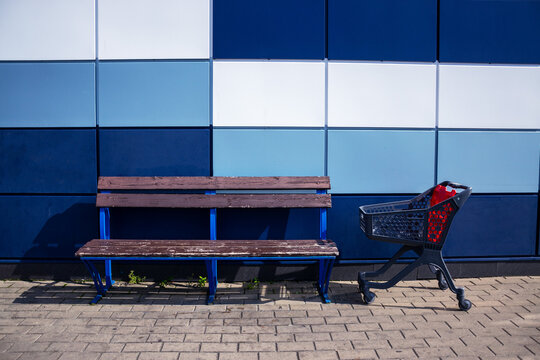 Empty plastic shopping cart standing next to wooden bench by colorful geometric wall of supermarket outdoors with copy space for retail business