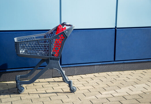 Empty grey plastic shopping cart with red details standing on paved sidewalk by blue wall outdoors with copy space for retail store concept