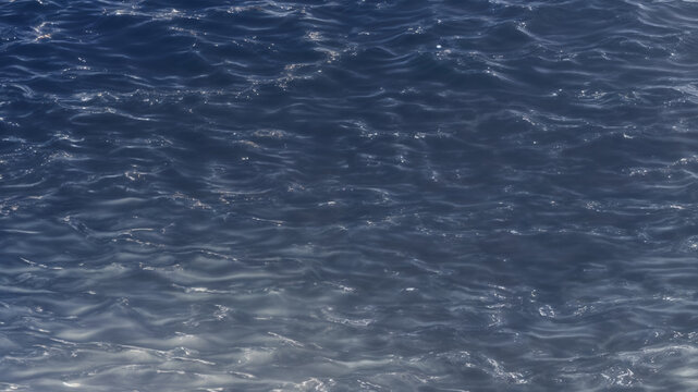 Close view of calm body of water with its surface shimmering under light to create a pattern of silver highlights and wave-like shapes with a deep blue background