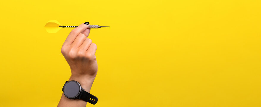 Close-up of a hand holding a dart against a bright yellow background, symbolizing focus, precision, and goal achievement. Modern watch visible on wrist, panoramic layout