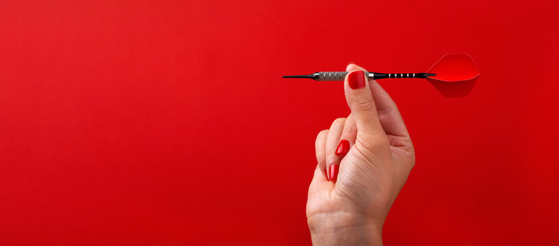 Close-up of a hand with red nails holding a red dart against a vibrant red background, ideal for concepts of precision, focus, and achievement, panoramic layout