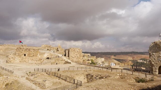 Wide exterior view of Kerak Castle in Jordan, showing medieval stone ruins, open courtyards and arched structures beneath dramatic storm clouds, with a Jordanian flag visible.