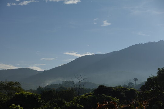view of multiple layers of hazy, dark mountains receding into the distance under a light blue sky with scattered wispy clouds. A faint mist or haze clings to the valleys and lower slopes of the distan
