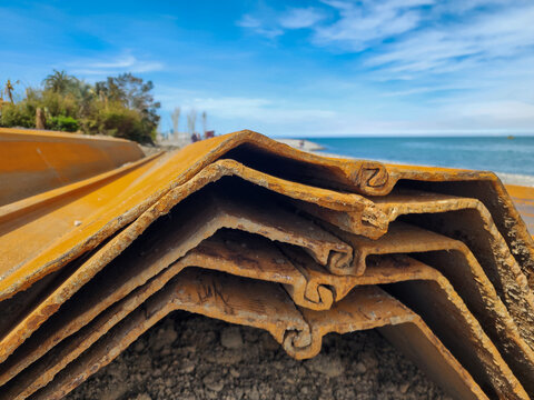 Rusty steel sheet piles stacked on a beach under a blue sky, preparing for construction work on coastal erosion control and shoreline protection against rising sea levels