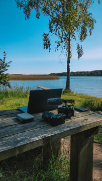 Laptop, camera, and microphone arranged on a rustic wooden table by a tranquil lake, representing a flexible workspace for digital nomads and creators working outdoors. Vertical photo