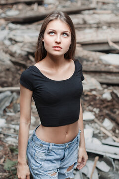Candid portrait of a slender young woman with long brown hair, looking upwards with a thoughtful expression, wearing a black crop top and ripped denim shorts amidst decaying urban ruins and rubble.
