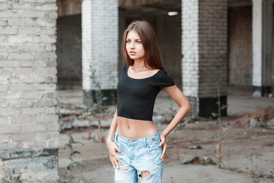 Attractive young woman with long brown hair, wearing a black crop top and ripped blue jeans, posing confidently amidst derelict concrete and brick pillars inside an abandoned, decaying industrial site