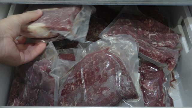 Close-up of a hand selecting vacuum-sealed frozen meat packages from a freezer, representing food storage, preservation and frozen meat.