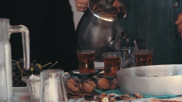 Person pouring hot water from kettle into traditional Turkish tea glasses on rustic table with walnuts