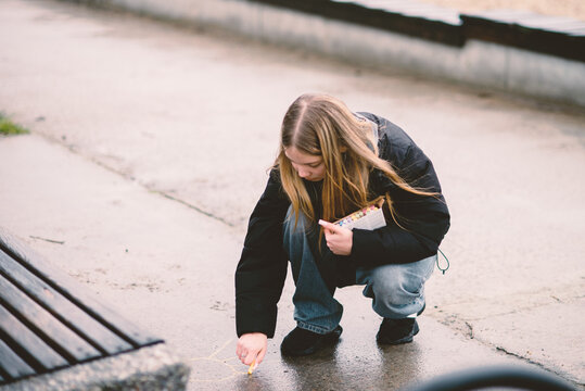 Young blonde girl squatting and drawing with colorful chalk on the pavement in a park