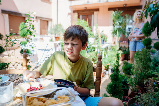 Young boy spreading berry jam on pancake during family breakfast in sunny courtyard in Spain