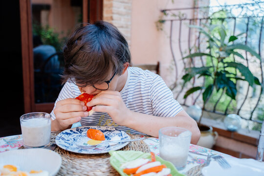 Teenage boy with Down syndrome eating tomato sandwich during relaxed family breakfast in Spain