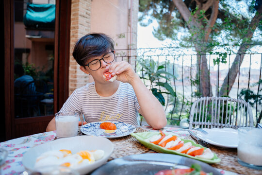 Teenage boy with Down syndrome enjoying tomato sandwich during everyday breakfast on terrace in Spain