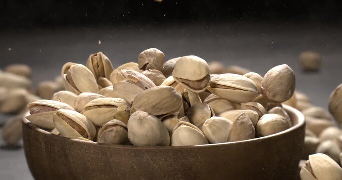 Wooden bowl filled with pistachios on dark wooden table, tasty salted nuts scattered around, healthy snack and natural food concept. Shot on super slow motion camera 1600 fps.