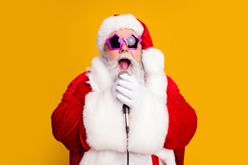 Naklejka premium Santa singing into a microphone wearing star sunglasses on a bright yellow background for a festive Christmas portrait and holiday party celebration