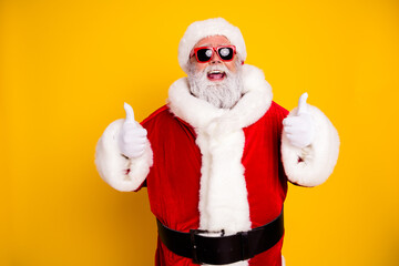 Naklejka premium Cheerful Santa Claus in sunglasses giving thumbs up on bright yellow background, festive Christmas portrait with red suit and white fur trim