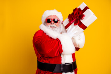 Naklejka premium Santa Claus holding a wrapped gift wearing red suit and sunglasses with bright yellow background, cheerful holiday portrait celebrating Christmas and gift giving