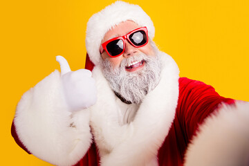 Naklejka premium Cheerful Santa Claus in red suit and sunglasses gives thumbs up selfie on yellow background celebrating Christmas holiday joy and festive winter costume portrait