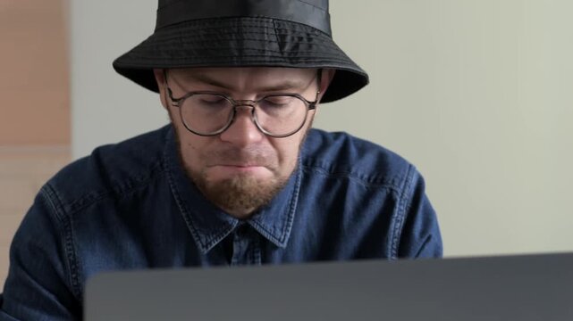 Indoors, young man sits at desk and leans forward, using both hands to adjust black hat while looking down at laptop, with man, young, adult, male detail, showing beard, stubble, glass, eyewear
