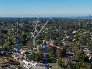 Obraz premium Construction cranes across a growing suburban Sydney district with established houses and distant skyline, illustrating redevelopment, housing growth and urban change
