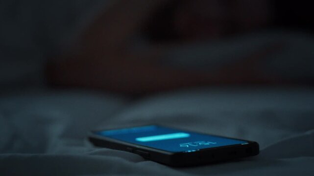 Unable to ignore her ringing phone a young woman wrapped in a blanket reaches for the glowing screen while half asleep. Dark bedroom. Theme of technology disturbing sleep and modern rest