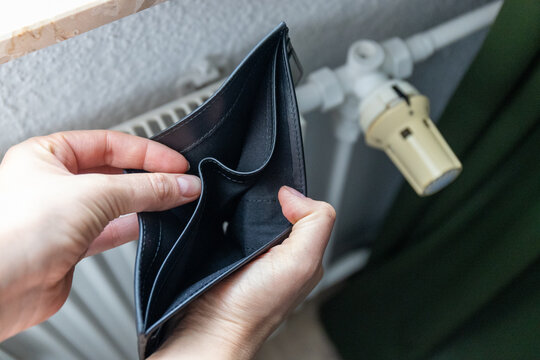 Hands holding an empty wallet in front of a heating radiator. Conceptual image of high energy bills and expensive utility costs.