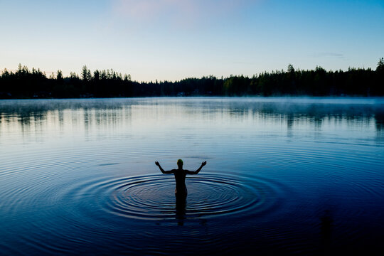 White person standing in calm lake water with ripples at misty forest