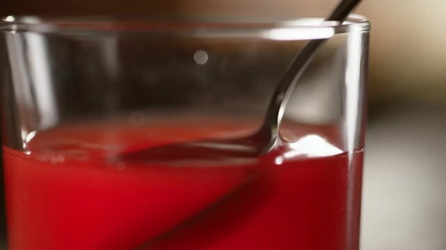 Red beverage stirred with a spoon in a clear glass in an indoor preparation setting. Beverage advertising, drink recipe videos, and bar and restaurant promotion.
