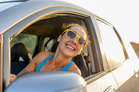 A cheerful woman wearing yellow sunglasses leans out of her car window during a sunny road trip.