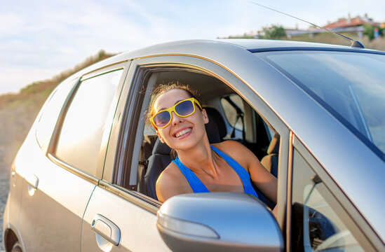 A cheerful woman with yellow sunglasses smiles out the open window of a silver car during a summer road trip.