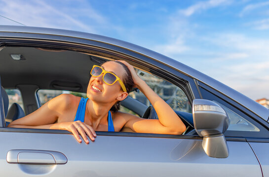 A cheerful woman in sunglasses looks out the open window of a silver car under a clear blue sky.
