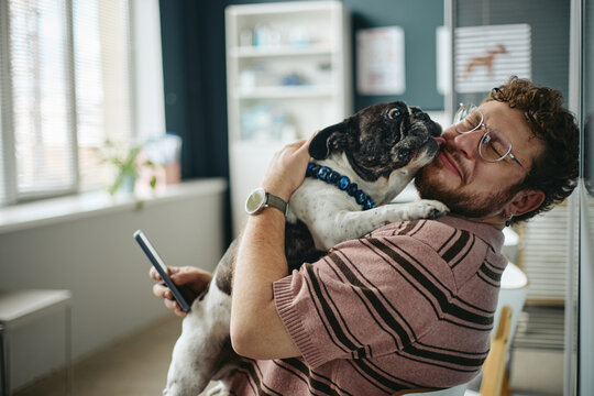 Young adult Caucasian man holding smartphone and hugging French bulldog licking his face in vet clinic. Commercial visual concept for pet care services and mobile veterinary apps