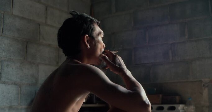 Tattooed man with earring smokes a cigarette, gazing upward against a weathered concrete wall in dim, introspective light.