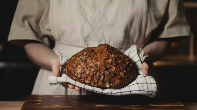 Close-up of baker in a rustic linen apron holding freshly baked artisan bread topped with pumpkin seeds on a checkered towel. Homemade loaf for bakery promos, organic product and healthy food concept.