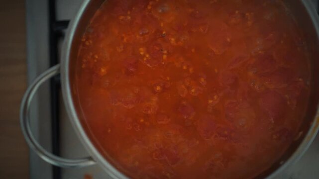 Overhead close-up of boiling tomato sauce in a metal pot. Active bubbles and tomato pulp pieces visible. Theme of traditional homemade cooking and natural pizza sauce preparation