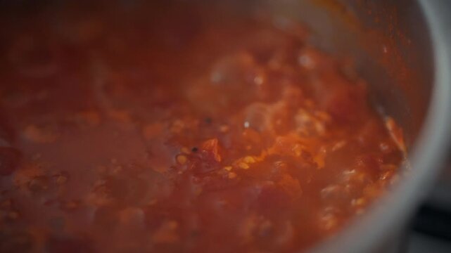 Top view of a metal pot with boiling tomato sauce on the stove. Tomato pulp and bubbles fill the surface. Concept of homemade organic sauce preparation and natural food cooking