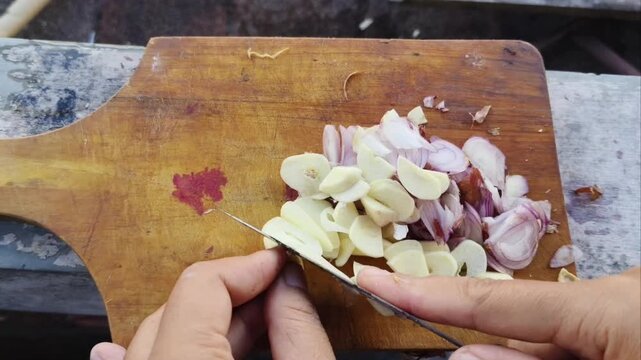 Top-down view of hands slicing fresh shallots and garlic cloves on a wooden cutting board, preparing ingredients for cooking a delicious meal.
