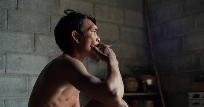 Tattooed man with earring smokes a cigarette, gazing upward against a weathered concrete wall in dim, introspective light.