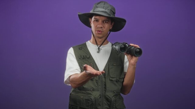 Man wearing safari hat and green vest holding binoculars with hand to mouth blowing kiss gesture in studio; playful curiosity.