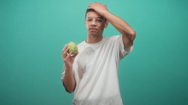 Man holding custard apple, hand on head with open mouth and raised eyebrow, mid sequence of surprised to thoughtful expressions, wearing glasses in studio; curiosity discovery.