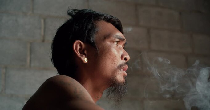Tattooed man with earring smokes a cigarette, gazing upward against a weathered concrete wall in dim, introspective light.
