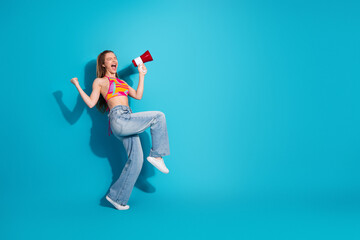 Excited young woman in vibrant outfit holding megaphone poses on blue background celebrating joy...