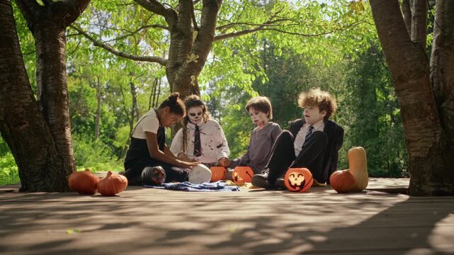 Multiethnic children and teenagers wearing Halloween costumes sitting on wooden deck with pumpkins, sharing autumn treats. Footage suitable for seasonal holiday campaigns and youth advertising