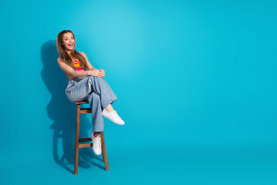 Young woman sitting on a stool with a cheerful smile against a vibrant blue background showcasing casual summer style