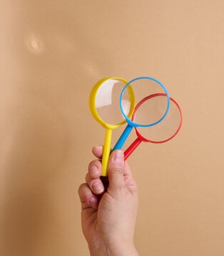 A person is holding three multicolored plastic magnifying glasses against a beige background.