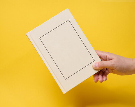 A person holding a closed light hardcover book with a frame on the cover against a yellow background