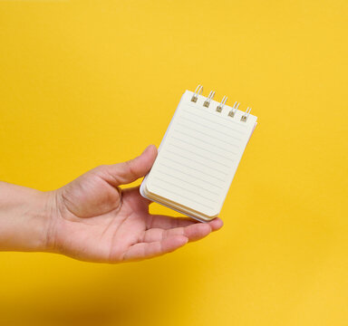 A person holding a small spiral-bound lined notepad on their palm against a bright yellow background