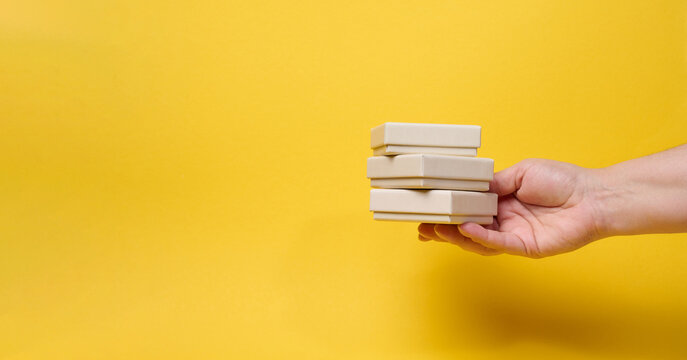 A woman's hand holds a stack of three small gift boxes on a bright yellow background.